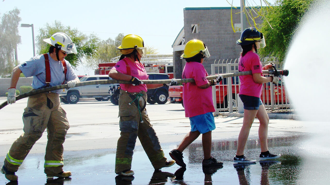 Young girls advancing a fire hose with firefighter helping from behind.